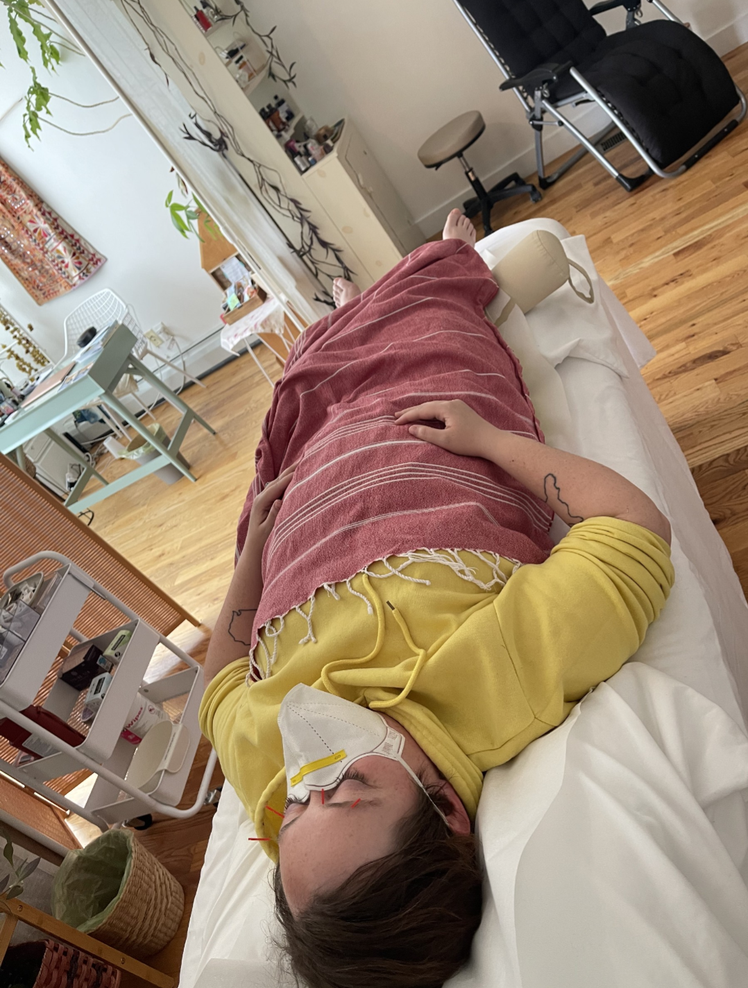 PJ is laying on a massage table in a well lit room. She's wearing a yellow hoodie, mask and a pink blanket