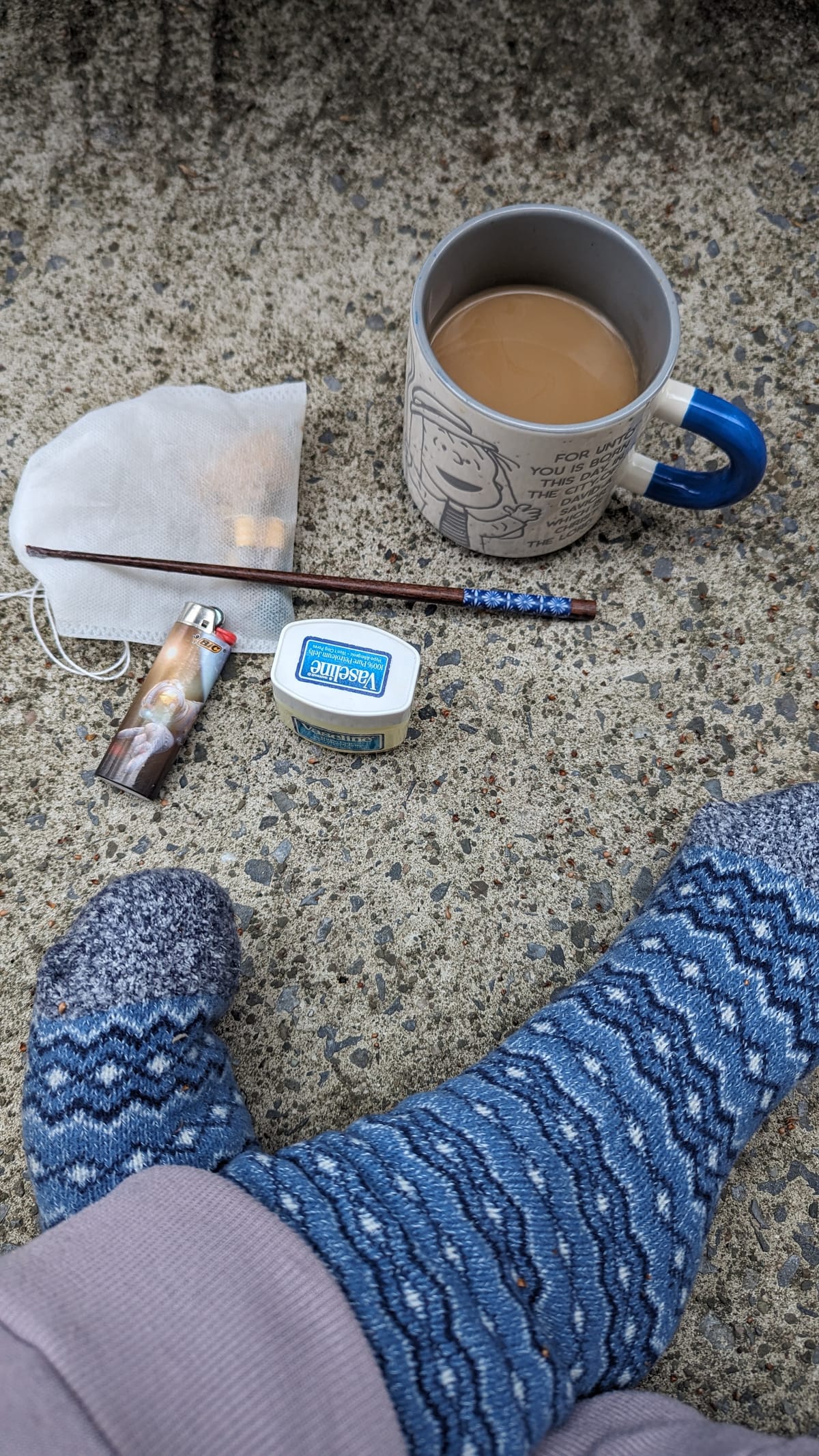 PJ's feet, in blue striped socks, against a stone patio background, there is a lighter, a container of salve, a chopstick, a bag containing mugwort, and a blue mug of coffee.