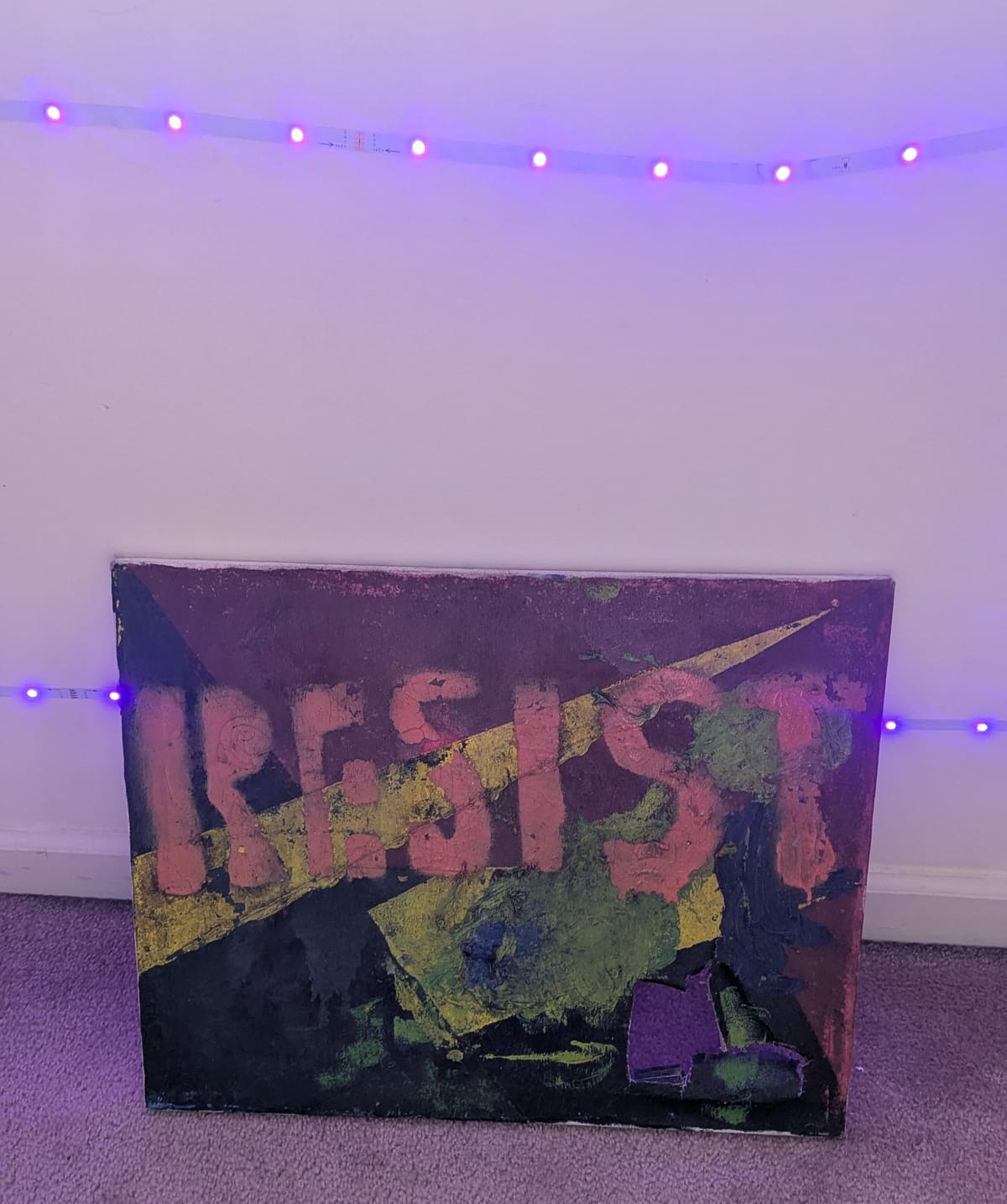 Purple LED lights outline a green, yellow and red painting with the word RESIST stenciled on, the canvas has a tear on the bottom right