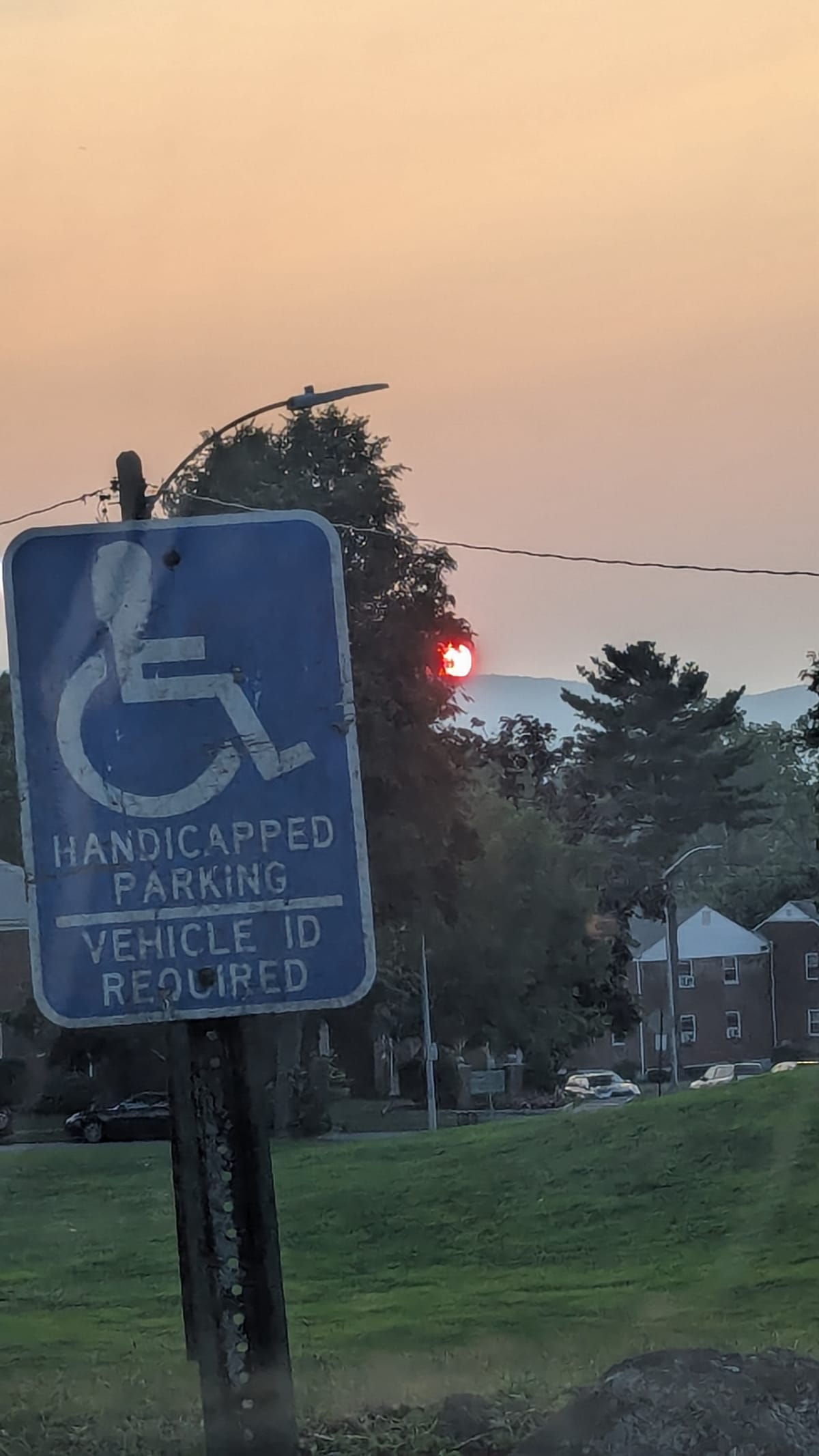 Close up of a Blue Disabled parking sign, against a backdrop of a grassy hill, apartments, and a bright red sunset in hazy orange sky
