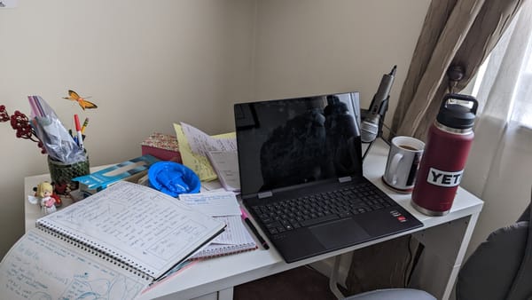 A messy white desk with some notebooks, an open laptop, a cup of coffee, a microphone and a maroon YETI water bottle