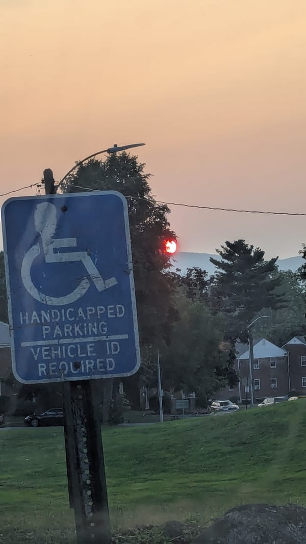Close up of a Blue Disabled parking sign, against a backdrop of a grassy hill, apartments, and a bright red sunset in hazy orange sky